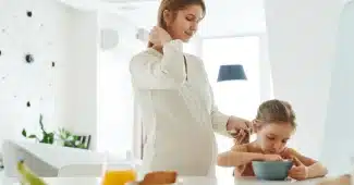 Mère prenant son petit-déjeuner à la cuisine le matin, avec un verre d'eau et une horloge en arrière-plan.