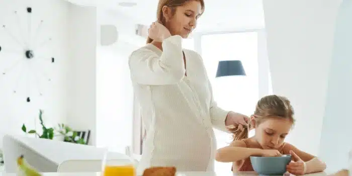 Mère prenant son petit-déjeuner à la cuisine le matin, avec un verre d'eau et une horloge en arrière-plan.