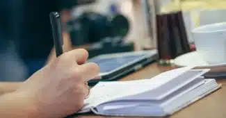 Carnet ouvert, tasse et mains écrivant une liste près d'une fenêtre en plein jour