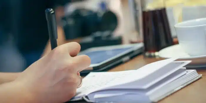 Carnet ouvert, tasse et mains écrivant une liste près d'une fenêtre en plein jour